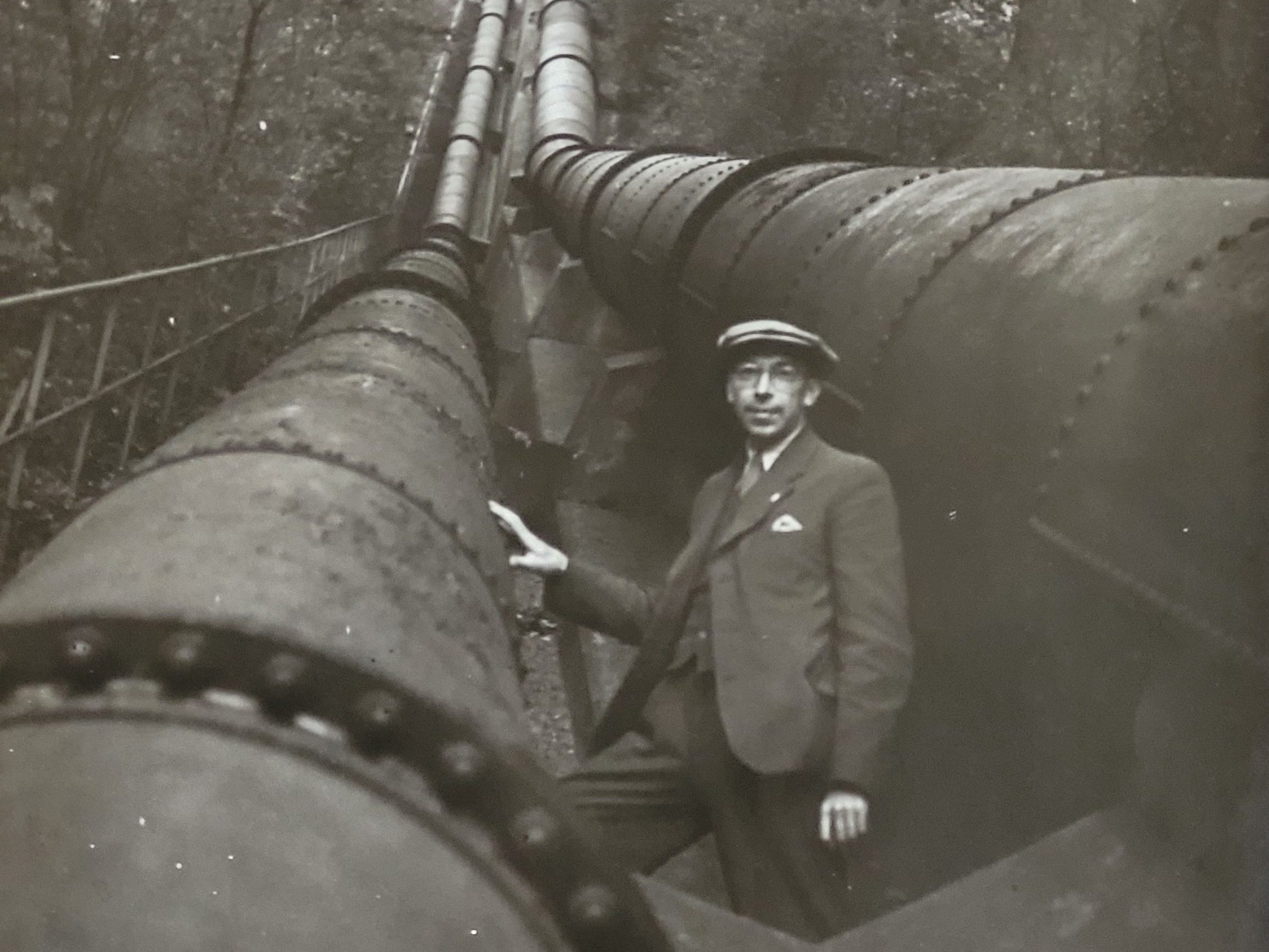 Schwarz-weiß-Foto aus den 1920er Jahren: Junger Mann mit Schiebermütze steht zwischen großen und sehr dicken Rohren - vermutlich Wasserleitungen.